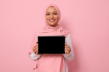 Arab Muslim beautiful woman wearing pink hijab, smiling toothy smile looking at camera, standing against pink background with copy space and showing the empty blank screen of a digital tablet