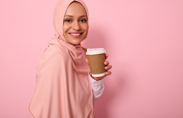 Close-up. Arabic Muslim woman with covered head in hijab holds disposable cardboard takeaway cup, smiles toothy smile, looking at camera, standing three quarters against colored background, copy space