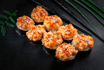 A set of spicy sushi rolls with baked salmon and teriyaki sauce. Maki rolls with nori on a black stone background with chopsticks