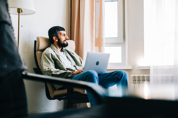 Caucasian man freelancer working at home, sitting on armchair and using laptop computer.