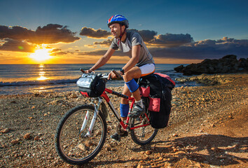 An athletic man in a bicycle uniform and a helmet rides a mountain bike with a large backpack on the trunk along the sandy shores of the Mediterranean Sea during a fantastic sunset. Cyprus