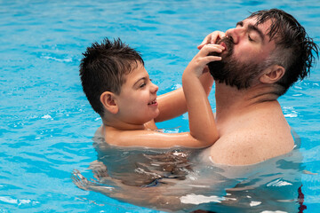 child with disability laughs and plays in his father's arms in a swimming pool - obrazy, fototapety, plakaty