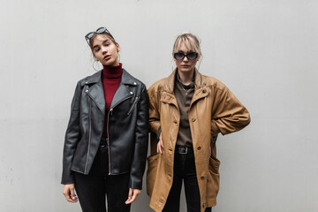 two beautiful teenage model girls in fashionable leather jacket with knitted sweater and jeans with sunglasses are posing near the gray wall on the street