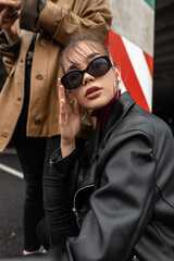 Trendy young model woman in fashion sunglasses in black leather jacket sits and posing on the street