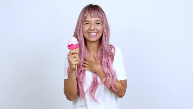 Young woman with pink hair holding a cornet ice cream smiling a lot while covering mouth over isolated background