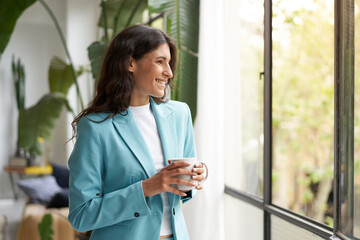 Young smiling business woman satisfied with a job well done relaxing with her morning coffee or tea, looking out the window. Beautiful latina woman celebrating successes in the office. - obrazy, fototapety, plakaty