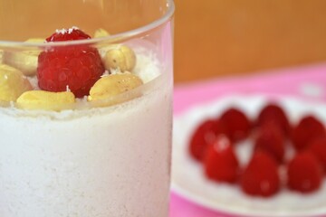 Close-up of coconut milk jelly in a glass decorated with cashews and raspberries and a plate of raspberries behind on a pink cloth with white polka dots
