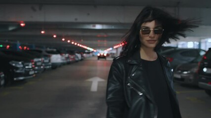 Following beautiful happy brunette woman wearing black leather jacket and sunglasses walking along underground parking with parked cars turns to camera flirts and smiling. Urban style fashion - Powered by Adobe