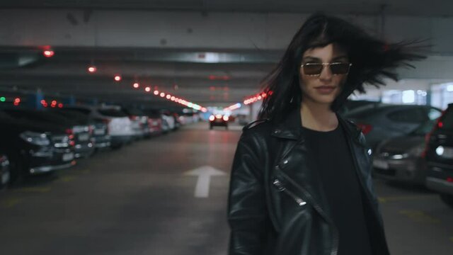 Following beautiful happy brunette woman wearing black leather jacket and sunglasses walking along underground parking with parked cars turns to camera flirts and smiling. Urban style fashion