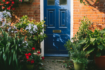 Fototapeta na wymiar Blue front door and brick walls facade of a Beautiful Cottage with many various potted flowers. The Small garden in front of the house, gardening hobby, create a cozy space around
