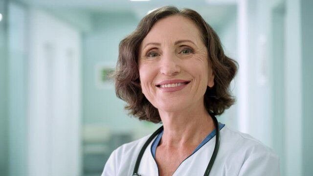 Portrait of happy smiling older senior female professional doctor physician pediatrician nurse wearing white robe standing in modern private clinic hospital looking at camera. Close up.
