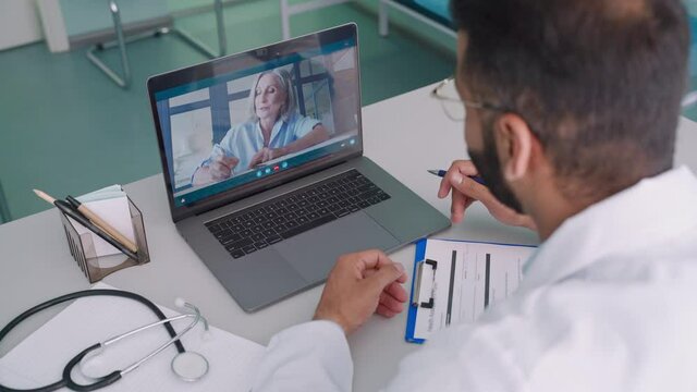 Videocall Chat With Female Older Patient And Indian Male Doctor In Modern Clinic Hospital Using Pc Laptop Computer, Consulting Online Remotely. Telemedicine Healthcare Concept. Over Shoulder View.