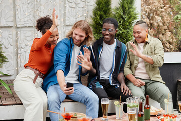 Diverse group of young people waving at camera via video chat while having fun during outdoor party at rooftop