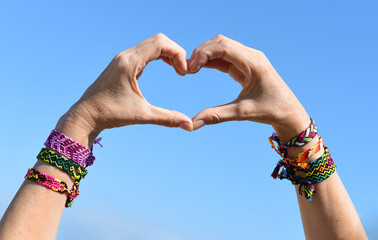 Hands making heart symbol on sky background. friendship bracelets on wrists - obrazy, fototapety, plakaty