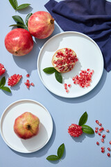 Shot of white plates of whole and sliced pomegranates, leaves and linen napkins on blue table. Eat clean. Dessert fruit. Topview 