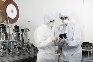 male engineers wearing personal protective equipment uniform(PPE) using tablet and checking medical face mask beside machine in laboratory