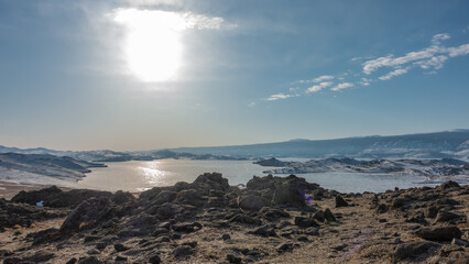 Obraz premium The frozen lake is surrounded by snow-covered hills. The sun is shining in the blue sky. Glare on the ice. In the foreground is dry earth and rocks. Baikal