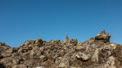 A fragment of picturesque granite rock against the blue sky. Cracks between boulders, dry grass on the slope. A pyramid of stones is built at the top. Siberia