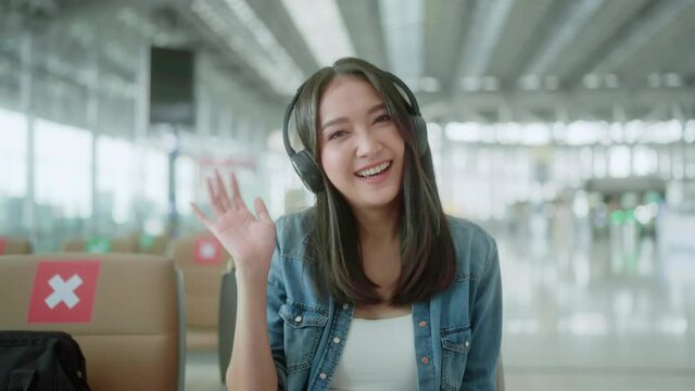 travel vacation concept,pov asian female woman wear headphone hand use laptop hand wave greeting to camera with cheerful and happiness,asian female sit wating at wating area in airport terminal