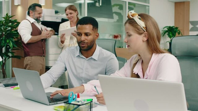 A blonde woman is helping out her colleague at the office with his work, they re both very happy while in the back two co-workers are conversing