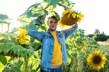 A young smiling man in a denim jacket and a yellow T-shirt stands under a huge sunflower on a sunny...