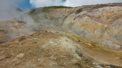 Geothermal zone on a mountain slope. The soil is covered with yellow-orange sulfur deposits. Steam and smoke rise from the fumaroles into the blue sky. Kamchatka