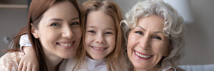Horizontal banner photo bonding loving three female generations family posing together, showing sweet tender feelings. Sailing adorable little kid girl cuddling beautiful mommy and old granny at home.