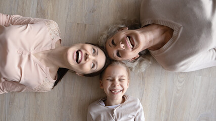 Overjoyed sincere laughing three female generations family lying on warm wooden heated floor, top...