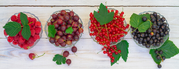 Garden berries in glass plates on a white wooden table: red currant, black currant, raspberry, gooseberry