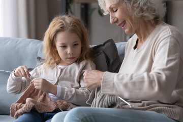 Smiling middle aged mature grandmother teaching adorable small child girl knitting stuff with...