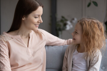 Smiling bonding young mother or nanny enjoying sincere conversation with cute small kid daughter, talking speaking communicating sitting on comfortable sofa in living room, trustful relations concept.