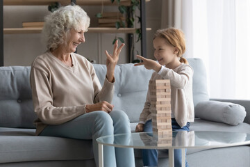 Laughing older granny deciding who will start game with smart little preschool kid girl, playing board funny development game sitting on sofa in living room, entertaining involved in domestic activity