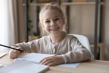 Portrait of smiling little pre teen kid girl learning, preparing for examination, enjoying handwriting in copybook doing school assignments, studying alone sitting at table at home, education concept.