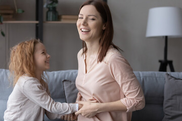 Joyful sincere small child girl tickling laughing young mother, having fun together in living room....
