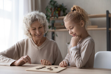 Caring middle aged old grandmother playing draughts with adorable little child girl, sitting...