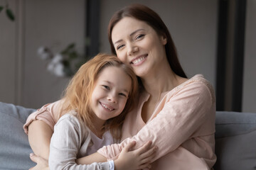 Portrait of affectionate young woman cuddling smiling little preschool child daughter, showing tender feelings at home. Joyful two different generations family enjoying loving moment together indoors.