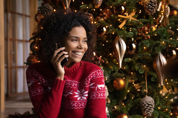 Smiling millennial African American woman near fir tree talk speak on cellphone on Christmas winter holidays. Happy biracial female client use smartphone have call online on New Year at home.