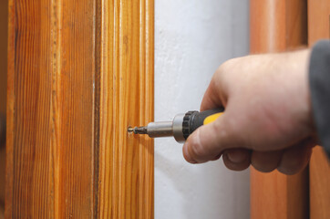 A man installs a wooden door trim. The hand screwdriver screws in the screw. Carpenter services. Construction industry. Indoors. Selective focus.