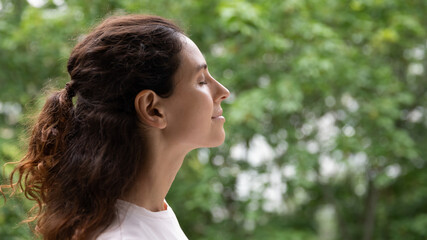 Profile close up face serene woman standing outdoor closed eyes breath fresh air feels motivated enjoy new day, green trees on background. No stress, filling with positive emotions, meditation concept - obrazy, fototapety, plakaty