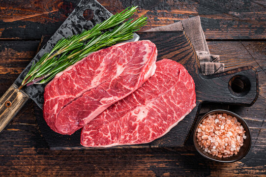 Uncooked Raw Shoulder Top Blade or flat iron beef meat steaks on a wooden butcher board with meat cleaver. Dark wooden background. Top View