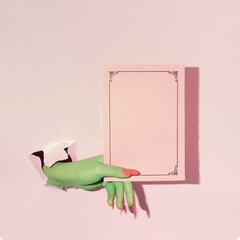 Creative layout with green painted hand holding pink paper copy space against pastel pink background. Halloween surreal idea. Minimal flat lay concept.
