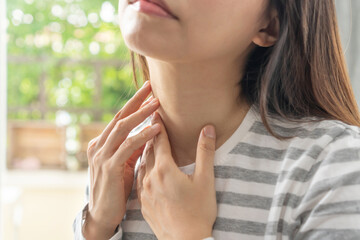 Unhappy Asian woman having sore throat, feeling sick. Ill girl suffering from painful swallowing, strong pain in throat, hand touching on her neck at home. Health problems concept.