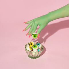 Creative layout with green painted hand holding green eyeball above dish with eyeballs against pastel pink background. Halloween surreal idea. Minimal concept.