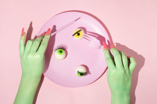 Creative layout with two green painted hands with fork and knife and green and yellow eyeball in pastel pink plate against pastel pink background. Halloween surreal idea. 