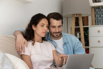 Millennial couple sit on comfy sofa with laptop buying goods on internet, discuss order, purchasing goods interior details on-line using web retail services. E-commerce buyers spouses family concept