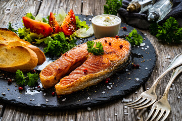Fried salmon steak with toasted bread and vegetable salad served on black plate on wooden table
