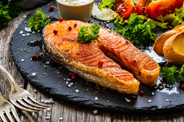 Fried salmon steak with toasted bread and vegetable salad served on black plate on wooden table
