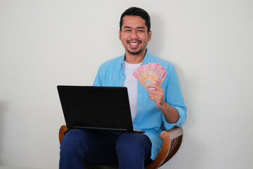 Adult Asian man sitting in a wooden chair while smiling happy showing his paper money