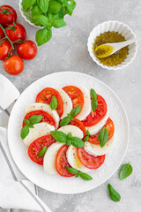 Classic caprese salad with mozzarella cheese, tomatoes and basil on a white plate on a gray concrete background. Top view, copy space.