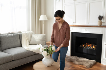 Cozy homey. Happy millennial indian woman young wife decorating luxury living room of family home put bunch of white roses on table before fireplace. Young mixed race female improving house interior - Powered by Adobe